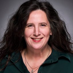 Smiling woman with long dark hair, wearing a dark green shirt, grey background.