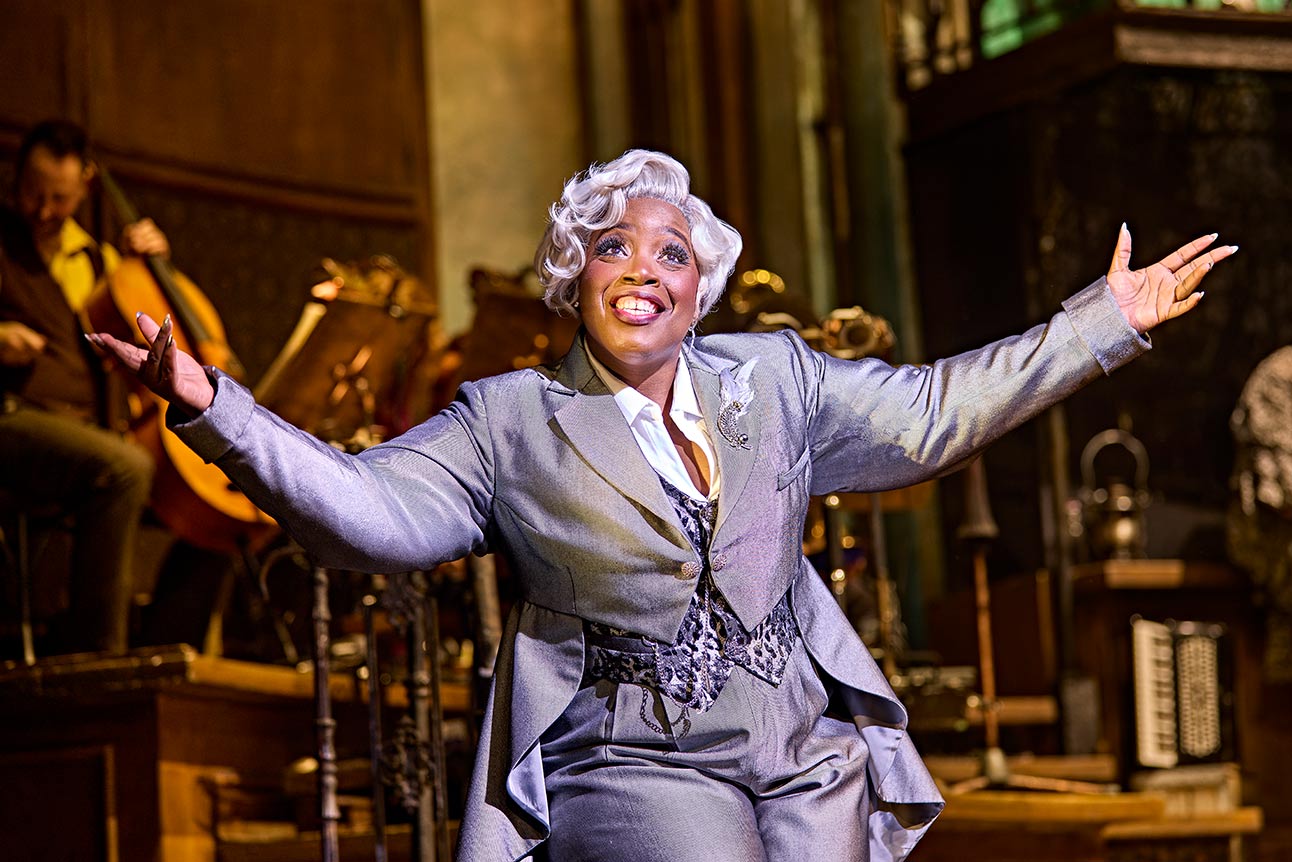 Malinda Parris performs energetically on stage in a grey suit, arms outstretched, with a joyful expression.