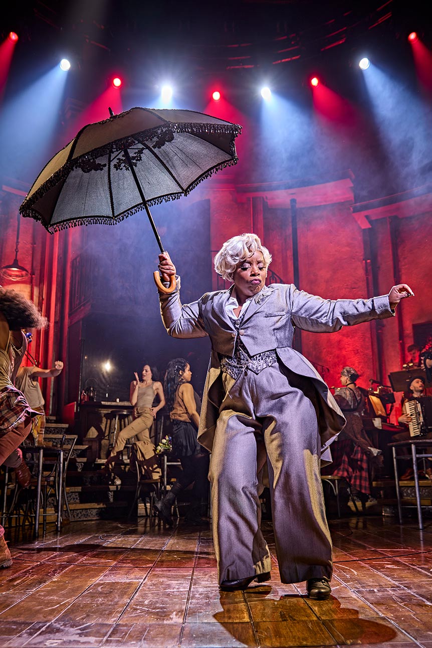 Malinda Parris performs energetically in "Hadestown," holding an umbrella on a vibrant, theatrical stage.