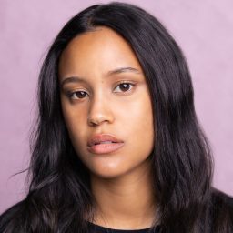 A woman with long black hair and a neutral expression against a light purple background.