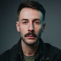 A man with short hair and a moustache, wearing a dark jacket, against a grey background.