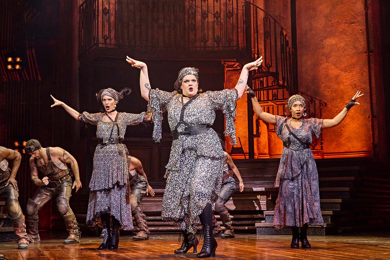 Melanie Bright, Spike Maxwell, and Lauran Rae perform energetically on stage in grey costumes, arms raised.