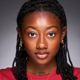 A person with braided hair, wearing a red top, looks directly at the camera against a grey background.