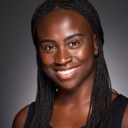 Smiling woman with braided hair, wearing a black top, against a dark grey background.