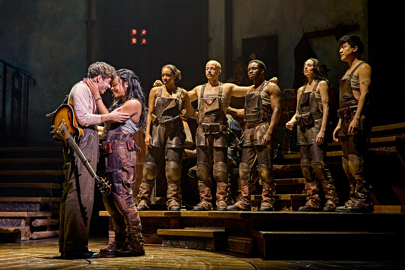 Nathan Sykes and Fayth Ifil embrace on stage with Hadestown cast in brown costumes behind.