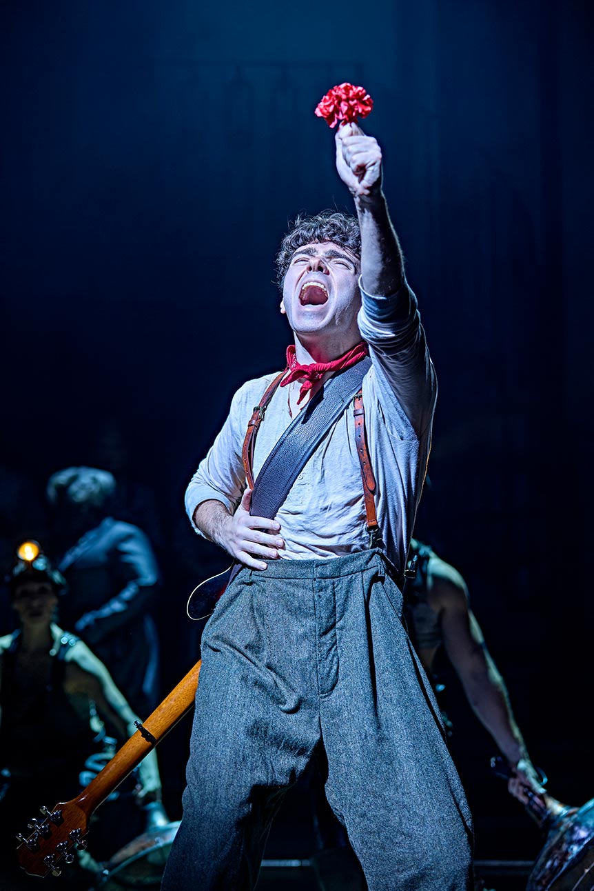 Nathan Sykes passionately sings on stage in "Hadestown," wearing a red scarf and suspenders.