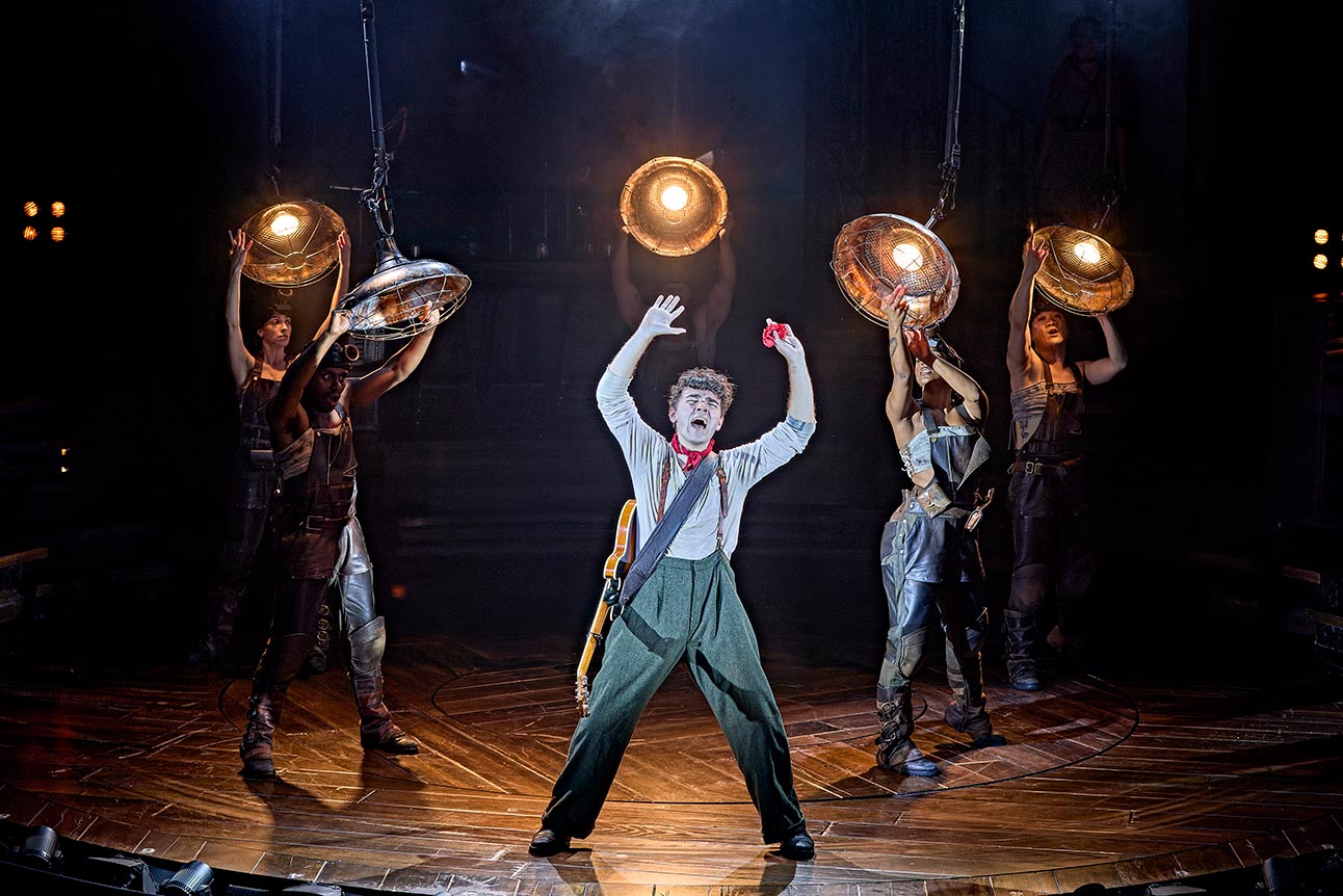 Performer in costume raises arms on stage, surrounded by three cast members holding lights.
