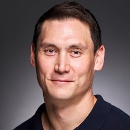 A man with short dark hair, wearing a dark shirt, smiles against a grey background.