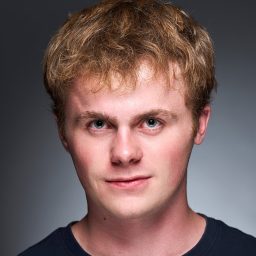 A person with short, light brown hair and a neutral expression against a grey background.