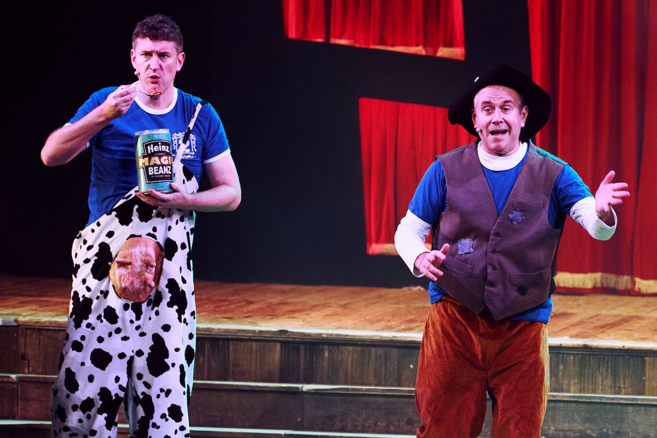 Two actors in colourful costumes perform energetically on stage at Wilton's Music Hall. one of them holds a tin of magic baked beans