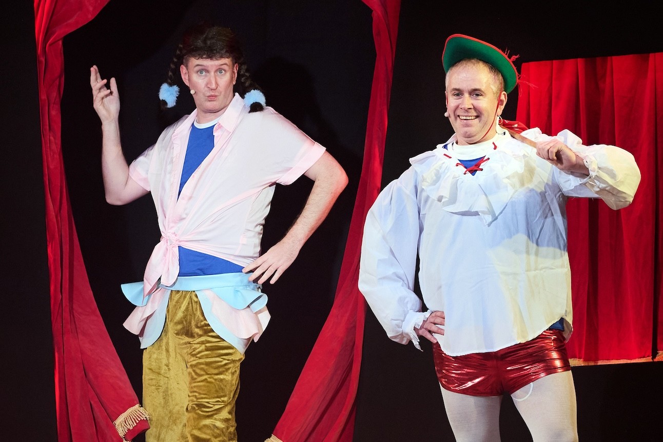 Alice in a blue dress holds a book, standing on stage with a red curtain backdrop.