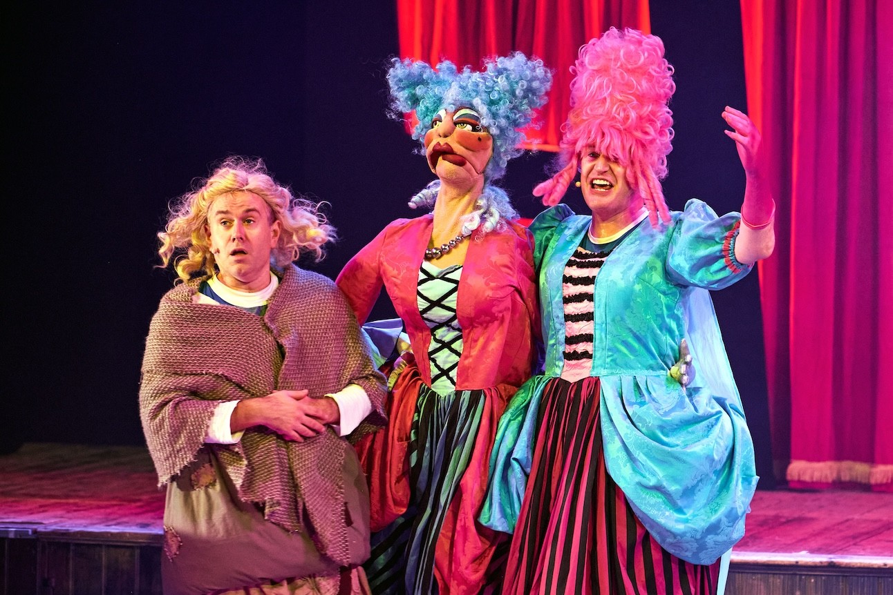 Two performers and a puppet in colourful costumes and wigs, standing on stage at Wilton's Music Hall.