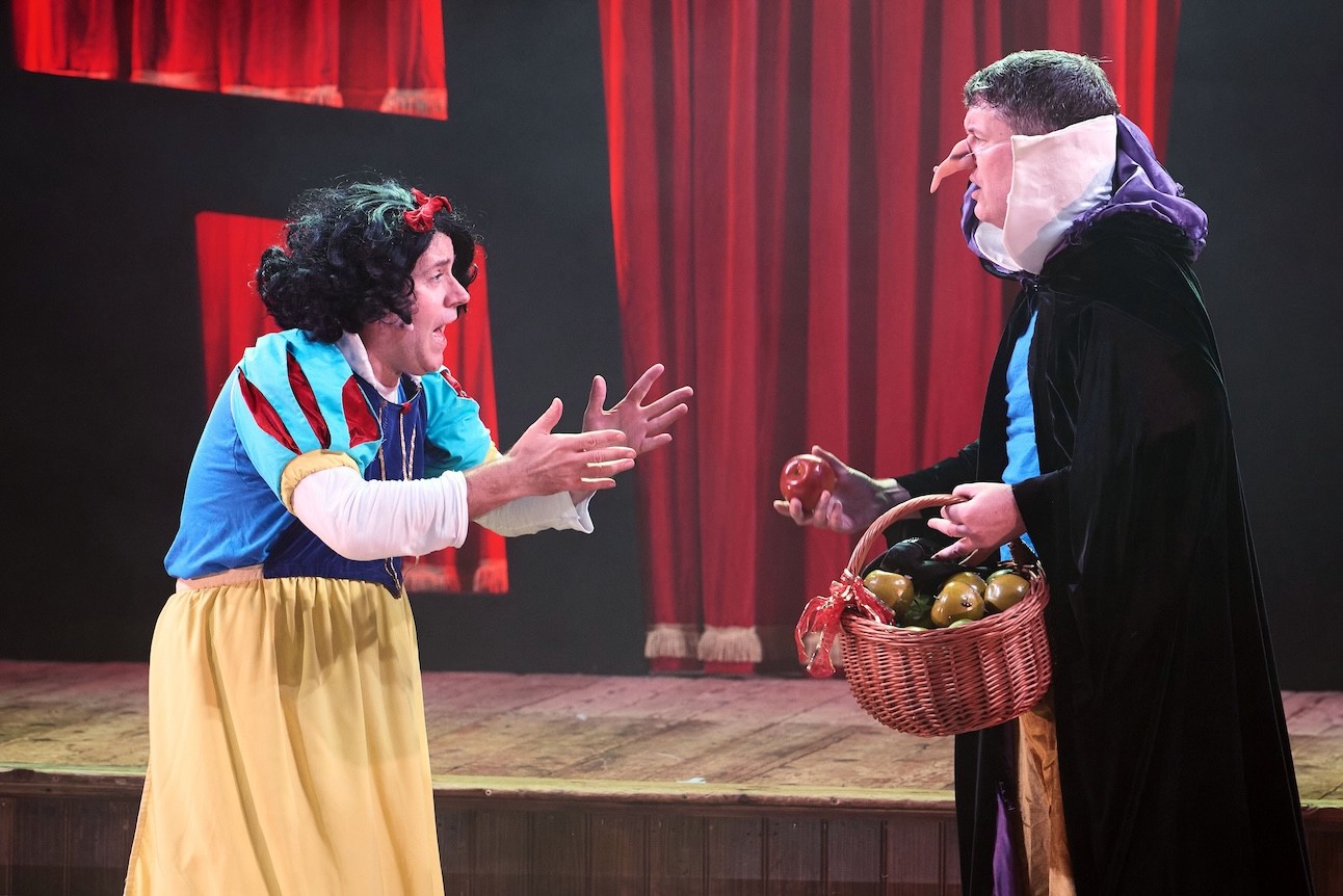 Snow White in a vibrant red and yellow dress stands smiling on stage at Wilton's Music Hall.