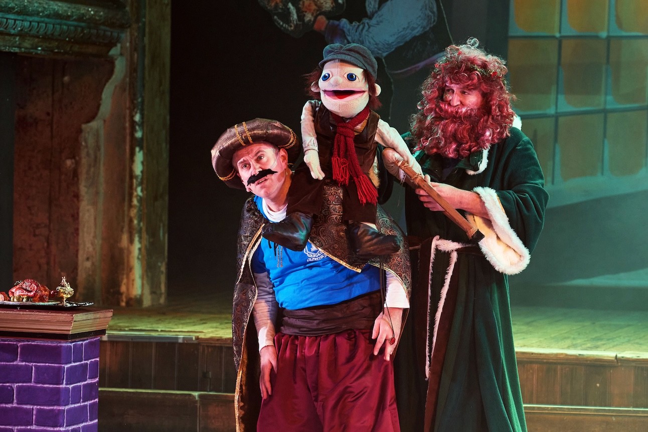 Two actors in costume, holding a puppet, stands on stage with colourful backdrop at Wilton's Music Hall.