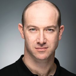 A man with a shaved head and a black shirt, looking directly at the camera.