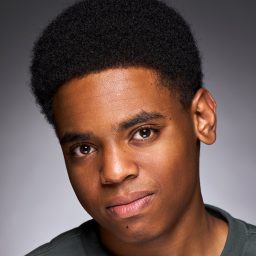 Young man with short curly hair, wearing a dark shirt, against a grey background.