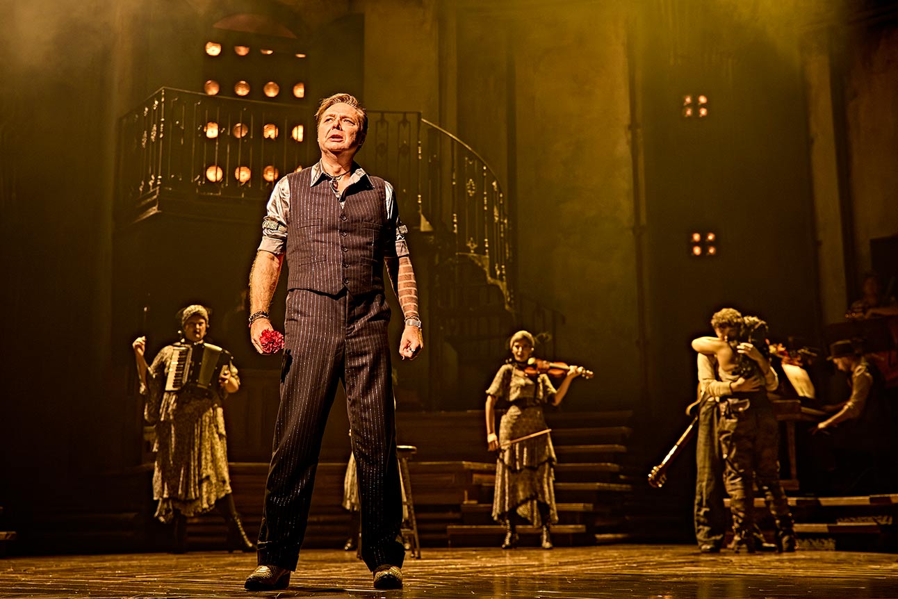 Shaun Dooley performs on stage with the cast of Hadestown, under warm yellow lighting.
