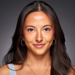 A woman with long brown hair, wearing hoop earrings and a blue top, smiles confidently.
