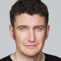 A man with short curly hair and blue eyes, wearing a black shirt, neutral background.
