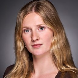 A person with long blonde hair looks directly at the camera against a grey background.
