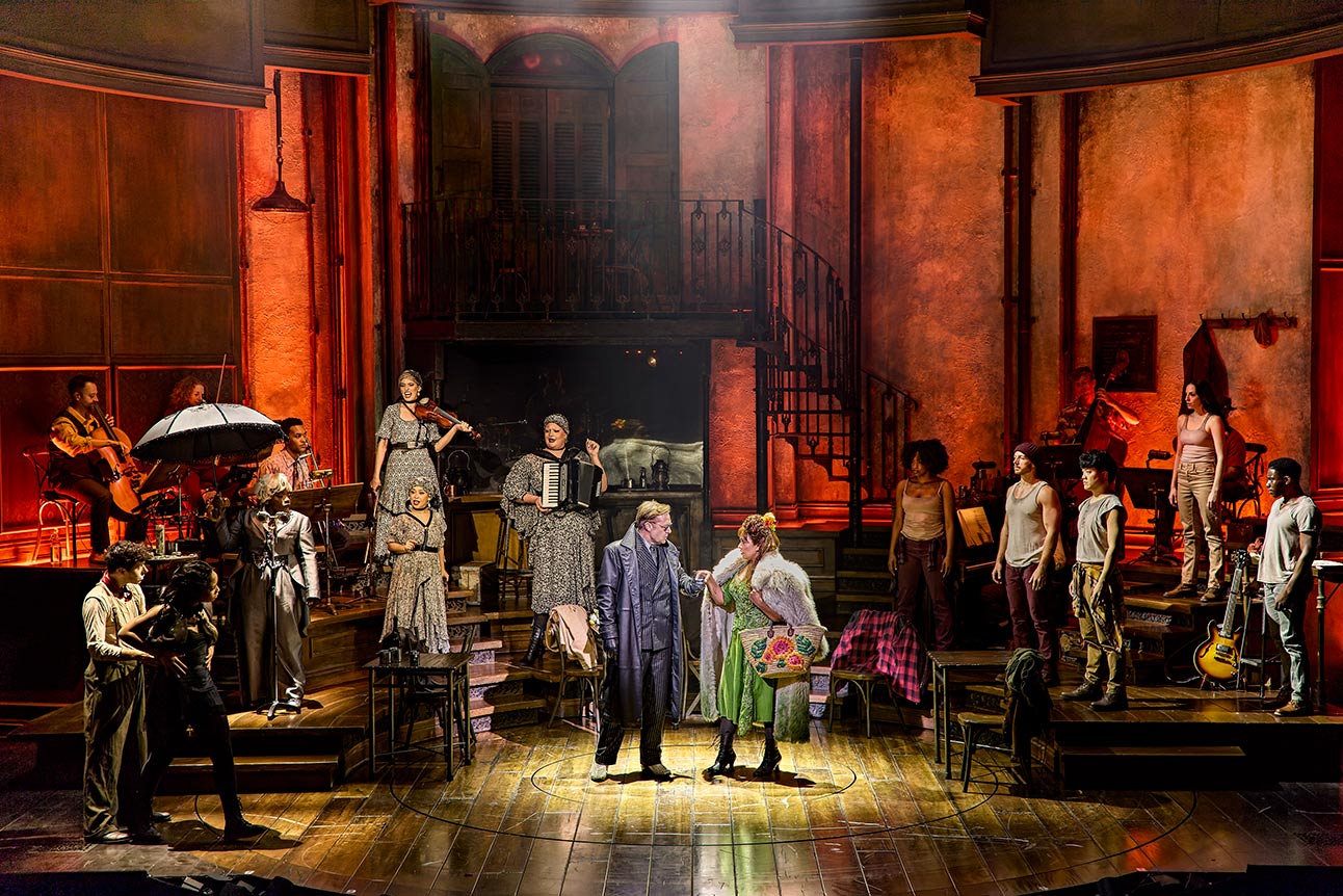 The cast of Hadestown performing on stage, with warm lighting and a rustic set design.