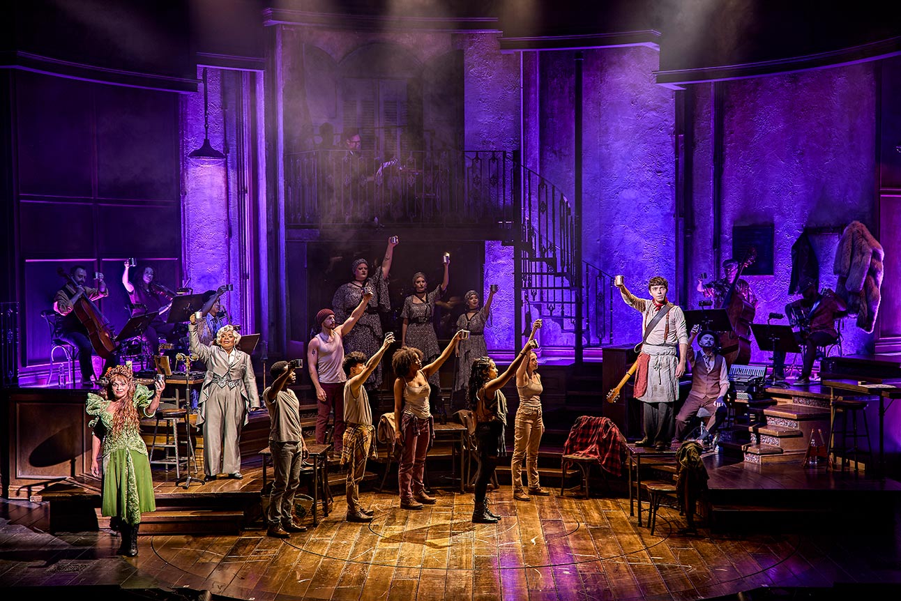 The cast of Hadestown performing on stage with purple lighting and raised hands.