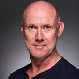 A bald man with a serious expression, wearing a dark shirt against a grey background.