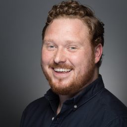 Smiling man with a beard and curly hair, wearing a dark shirt against a grey background.
