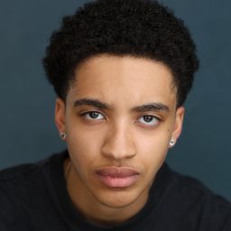 Young person with short curly hair, wearing earrings, against a plain blue background, looking directly ahead.
