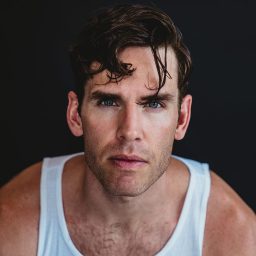 Man with short dark hair wearing a white vest top against a dark background, looking serious.