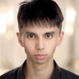 A person with short dark hair, wearing a black shirt, looks directly at the camera.
