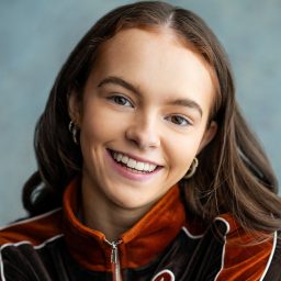 Smiling woman with long brown hair, wearing an orange and black jacket, against a grey background.