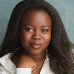 A woman with long dark hair, wearing a white shirt, poses against a grey background.