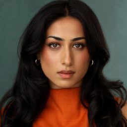 Woman with long dark hair in orange top, neutral expression, teal background.
