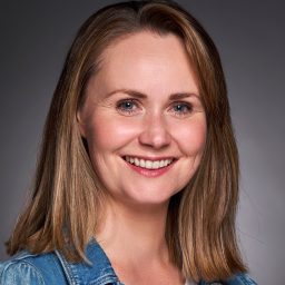 Smiling woman with shoulder-length hair, wearing a denim jacket against a grey background.
