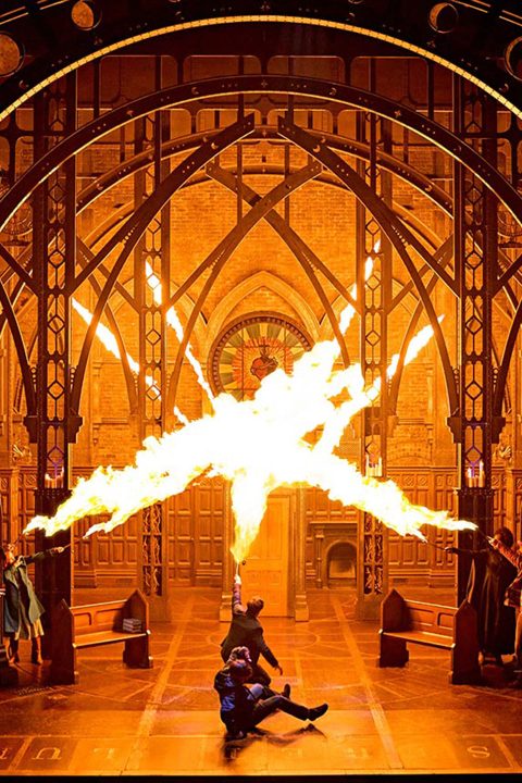 Dramatic stage scene with fiery explosion, gothic arches, and actors in dynamic poses, orange lighting.