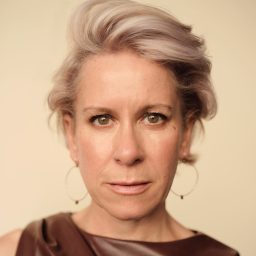 A woman with short, styled grey hair and a brown top, against a beige background.