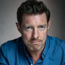 I don't know who this is. Man with short hair and beard, wearing a blue shirt, neutral background.