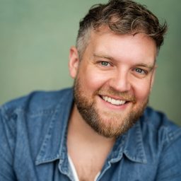 Smiling man with a beard in a denim shirt against a soft green background.