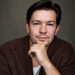 A man with dark hair, wearing a brown sweater, poses thoughtfully against a grey background.