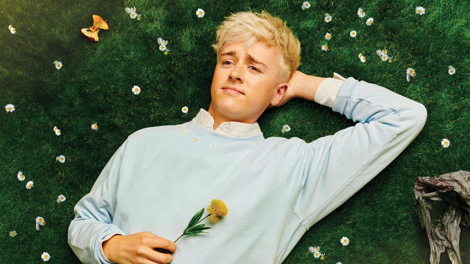 Blonde man in light blue jumper lies on grass, holding a yellow flower, daisies scattered around.