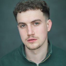 Young man with short curly hair, wearing a green top, against a grey background.