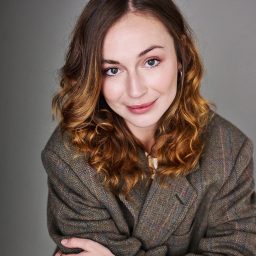 A woman with wavy brown hair wearing a grey blazer, smiling against a plain background.