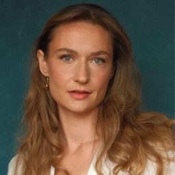 A woman with long brown hair and a white top against a dark teal background.