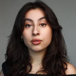 A woman with long dark hair and neutral expression against a plain grey background.
