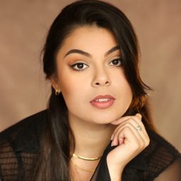 A woman with long dark hair poses thoughtfully, wearing a black top and gold necklace.