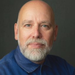Bald man with a beard wearing a blue shirt against a dark grey background.