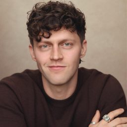 A man with curly hair wearing a dark sweater, smiling against a neutral background.