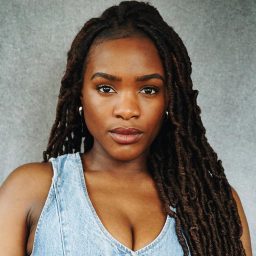 Woman with long braided hair wearing a denim top, neutral expression, grey background.