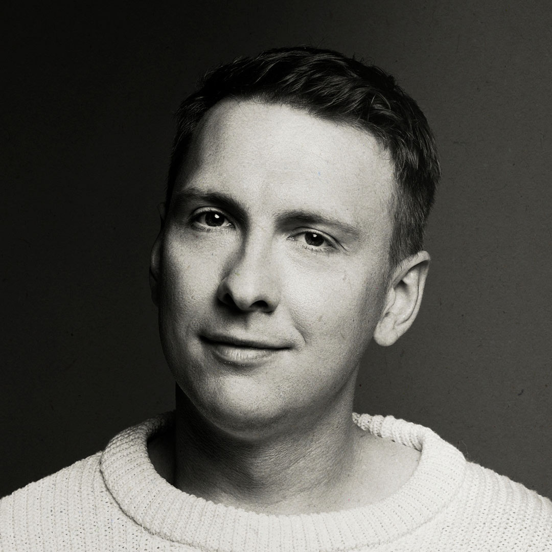 A person in a white jumper, smiling slightly, against a dark grey background.
